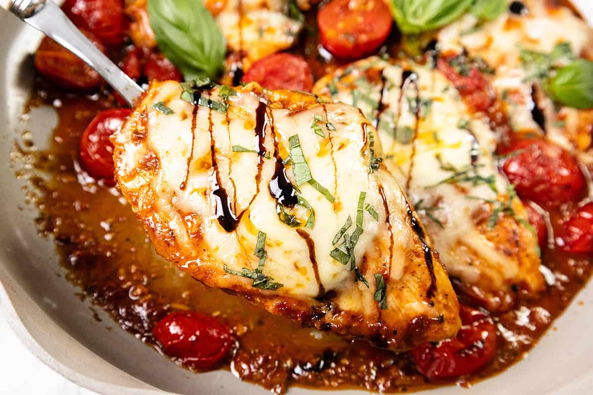 A close-up of Caprese Chicken: a cheesy baked chicken breast topped with fresh basil and a drizzle of balsamic glaze, served with roasted cherry tomatoes in a pan.