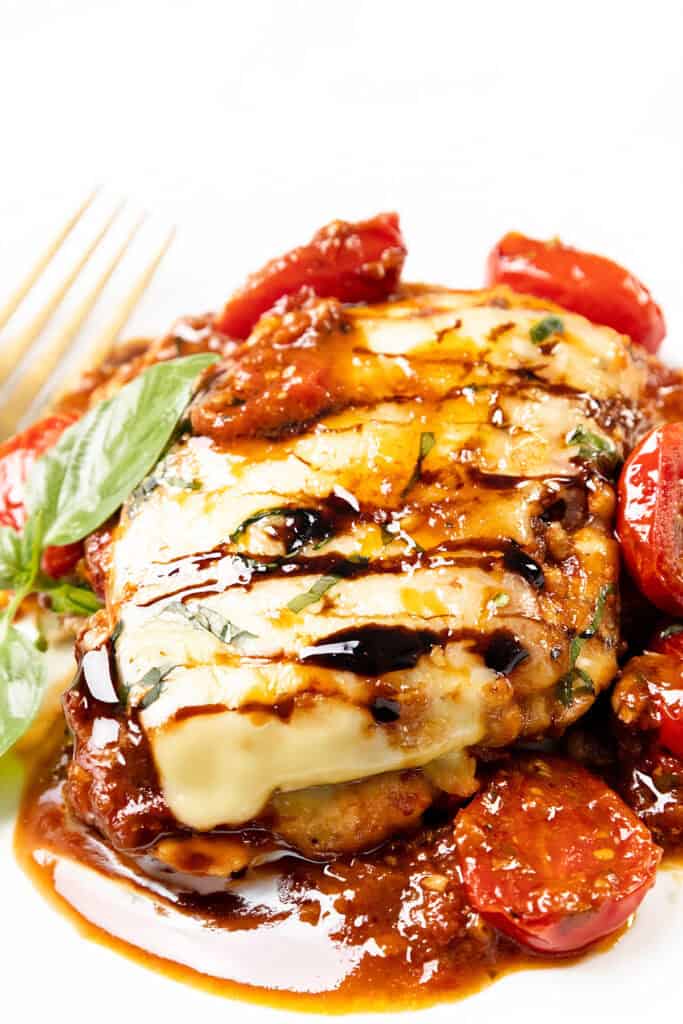 A close-up of Caprese Chicken parmesan topped with melted cheese, balsamic glaze, fresh basil, and roasted cherry tomatoes, with a fork in the background on a white plate.