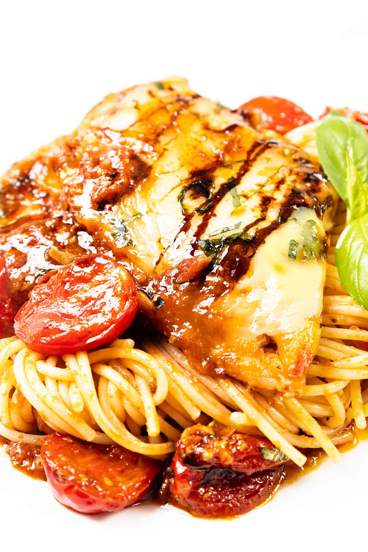 A plate of Caprese Chicken spaghetti topped with grilled chicken breast, melted cheese, tomato sauce, cherry tomatoes, and a drizzle of balsamic glaze, garnished with fresh basil.