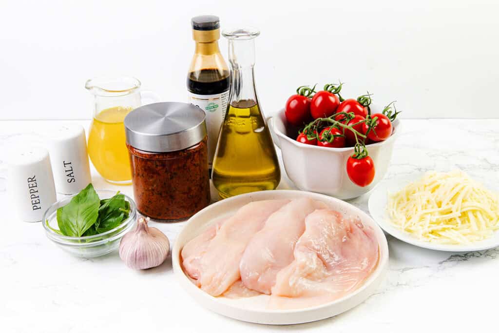 Several raw chicken breasts on a plate sit among fresh basil, garlic, cherry tomatoes, shredded cheese, oil, a jar of sauce, pepper, salt, broth, and soy sauce—perfect for crafting a flavorful Caprese Chicken on a white surface.