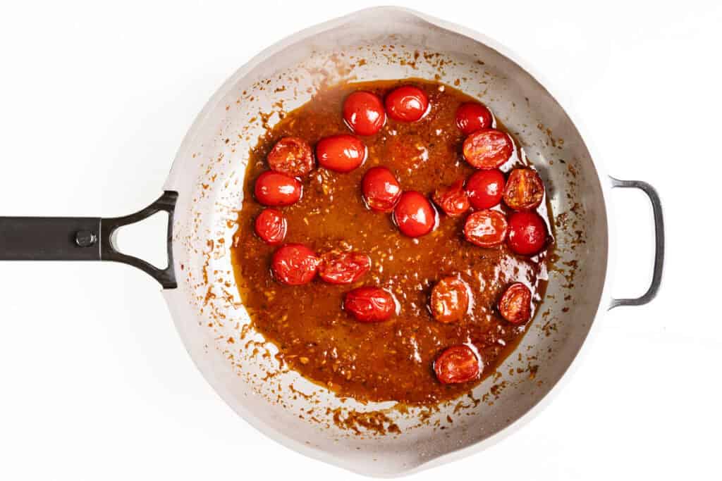 A skillet of Caprese Chicken with cherry tomatoes in a thick, reddish-brown sauce, some tomatoes whole and some halved, on a white background.