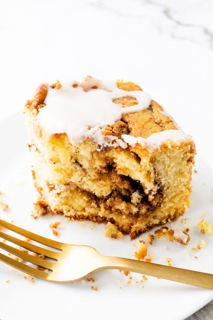 A slice of Cinnamon Bun Cake with white icing, partially eaten, sits on a white plate with a gold fork beside it.