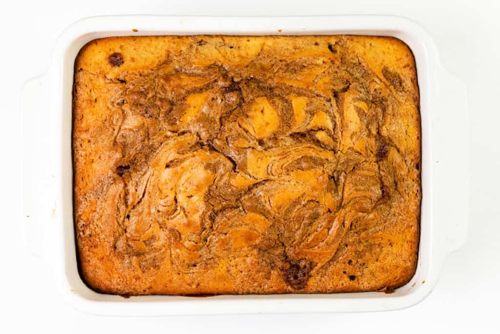 A rectangular baking dish filled with a golden-brown Cinnamon Roll Cake, featuring marbled swirls on the surface, viewed from above on a white background.