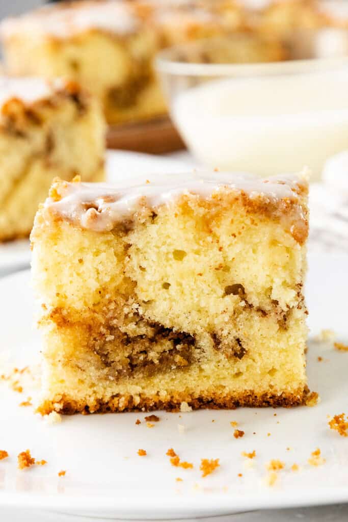 A close-up of a slice of Cinnamon Bun Cake with a light glaze on top, showing a moist, crumbly texture and a swirl of cinnamon in the center, served on a white plate.