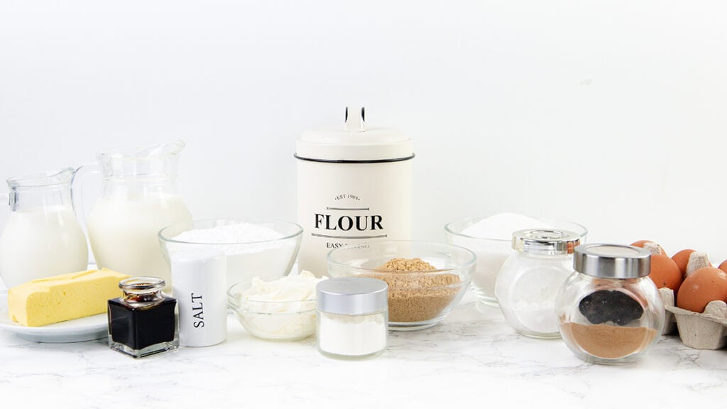 Various baking ingredients for a delicious Cinnamon Bun Cake, including flour, sugar, brown sugar, butter, eggs, milk, vanilla extract, salt, cocoa powder, and sour cream, are displayed in glass containers and bowls on a white countertop.