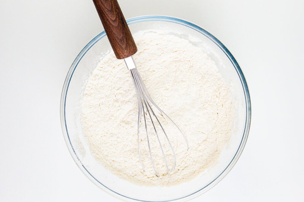A glass bowl filled with flour and a metal whisk with a wooden handle resting inside, ready for crafting a delicious Cinnamon Bun Cake on a white surface.