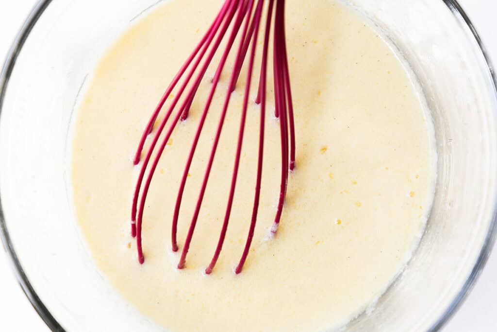 A red whisk mixing a pale yellow Cinnamon Bun Cake batter in a clear glass bowl, viewed from above.