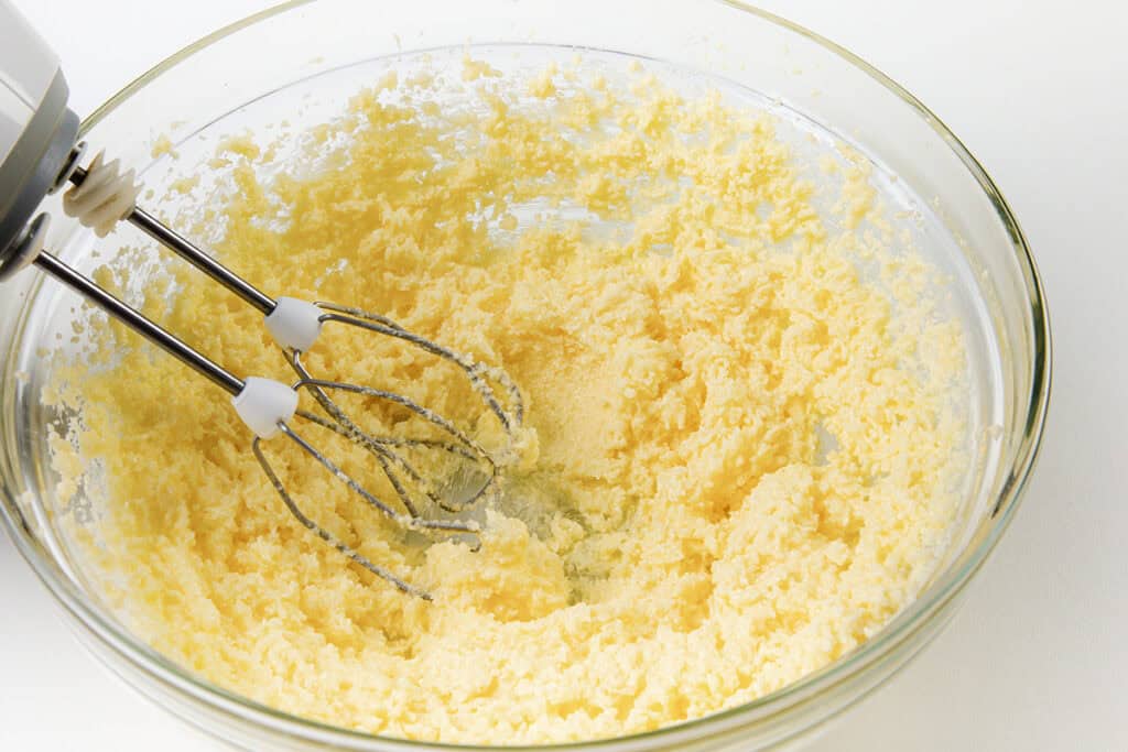 A hand mixer with two beaters sits in a glass bowl containing a pale, fluffy mixture for Cinnamon Bun Cake. The bowl is placed on a white surface, ready to create an irresistible treat.