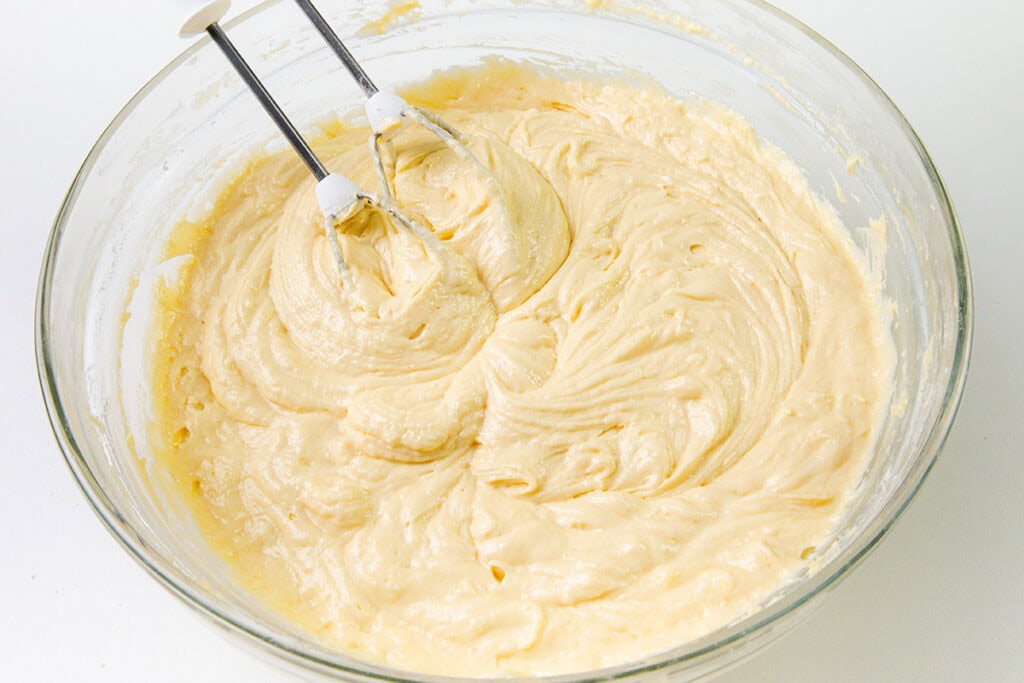 A glass bowl filled with creamy Cinnamon Bun Cake batter, with two metal beaters from an electric mixer resting in the mixture on a white surface.