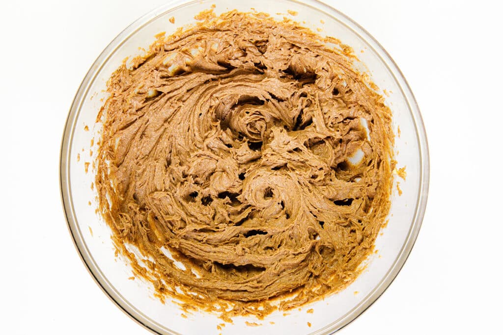 A glass bowl filled with light brown, creamy Cinnamon Bun Cake dough mixture, swirled and textured, sitting on a white background.