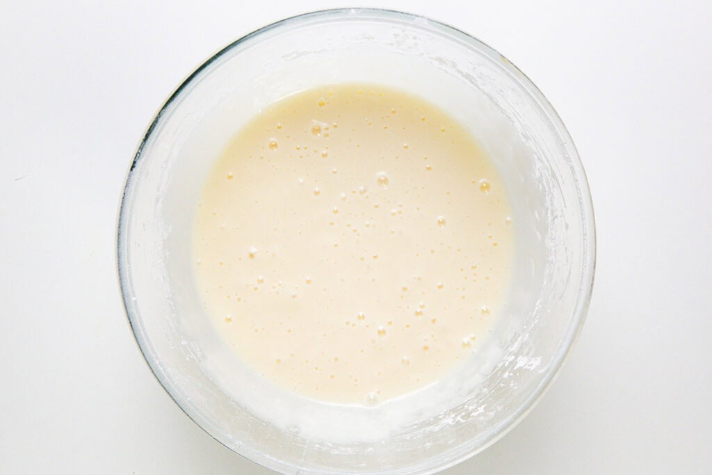 A glass bowl filled with smooth, pale Cinnamon Bun Cake batter sits on a white surface. Small bubbles are visible on the surface of the batter.