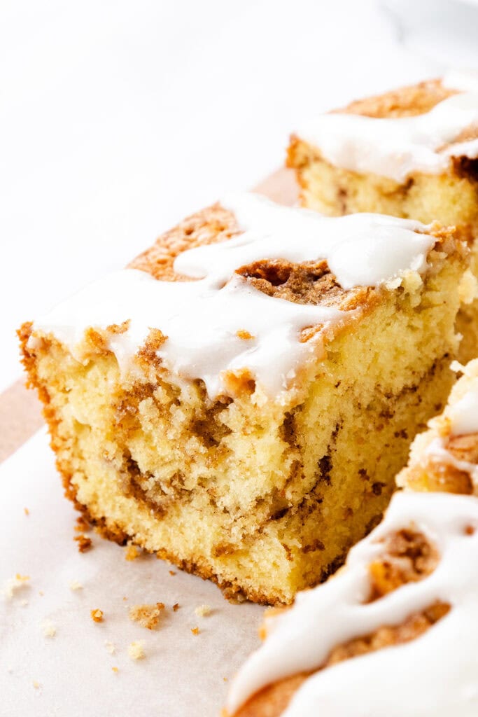 A close-up of a Cinnamon Bun Cake slice topped with white icing, showing its soft, moist texture and cinnamon layers inside. The cake sits on parchment paper.