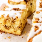 A close-up of sliced Cinnamon Bun Cake with a crumbly texture and swirls of cinnamon, topped with a white icing drizzle, displayed on parchment paper.