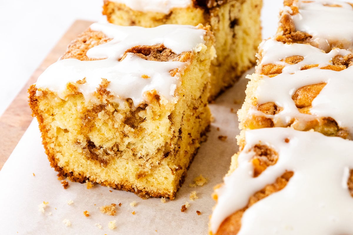 A close-up of sliced Cinnamon Bun Cake with a crumbly texture and swirls of cinnamon, topped with a white icing drizzle, displayed on parchment paper.