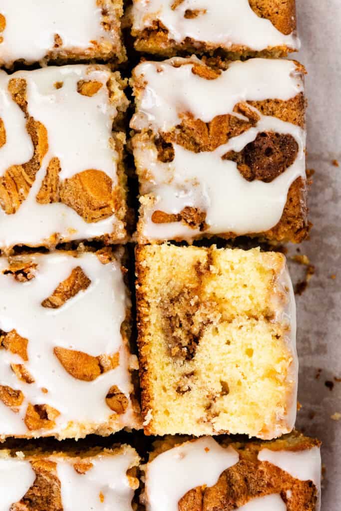 Close-up of sliced Cinnamon Bun Cake pieces topped with white icing, showing a moist, yellow crumb and visible cinnamon swirls, arranged in a grid on parchment paper.