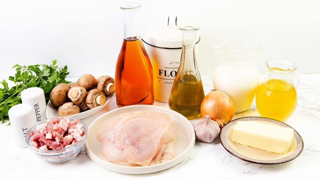 A variety of cooking ingredients on a white surface, perfect for making Creamy Chicken Marsala, including raw chicken breast, mushrooms, fresh parsley, pancetta cubes, onion, garlic, butter, olive oil, cream, flour, vinegar, salt and pepper shakers.