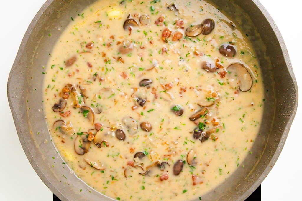Creamy mushroom sauce with chopped herbs and small pieces of bacon cooking in a large gray skillet, perfect for pairing with creamy marsala chicken. The sauce appears thick with visible mushroom slices and green herbs throughout.