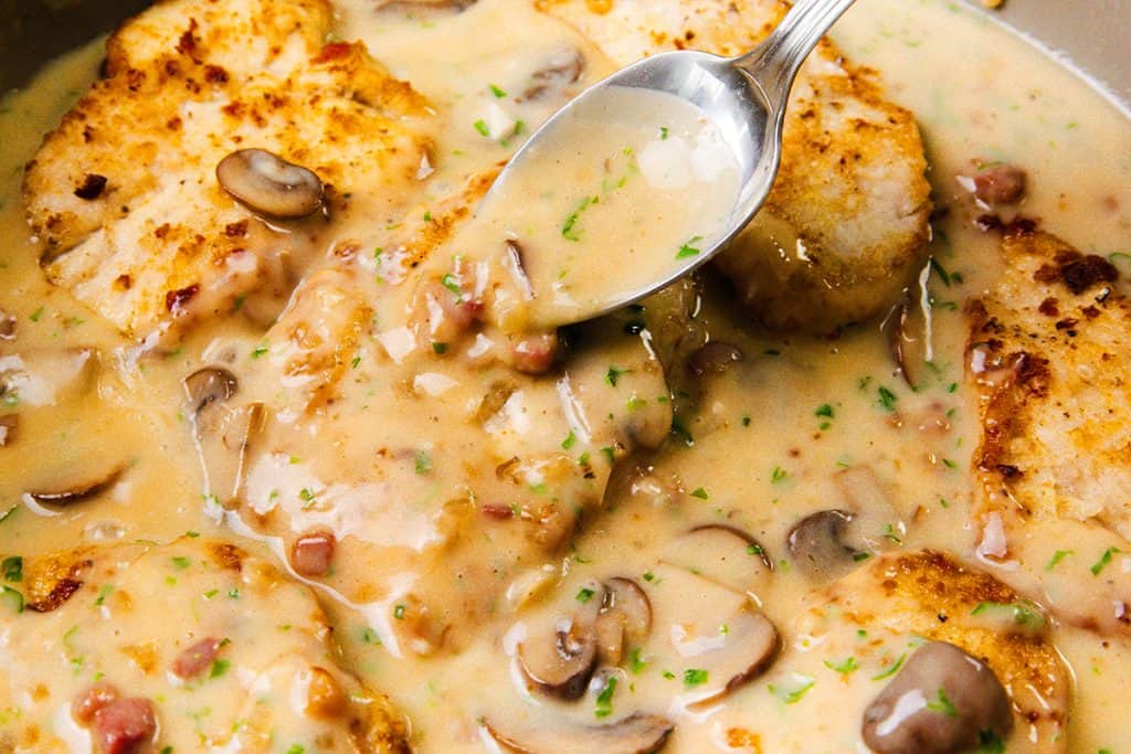 A close-up of Creamy Chicken Marsala cutlets in a luscious mushroom sauce, garnished with herbs. A spoon lifts the creamy sauce, revealing chopped mushrooms and bits of bacon.