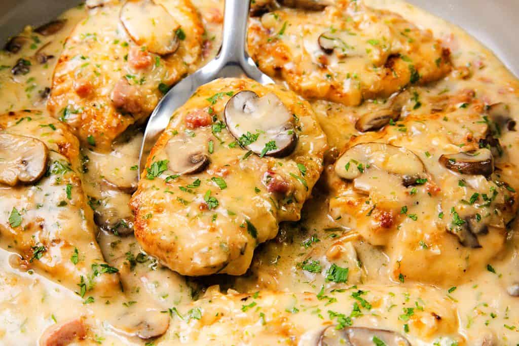 Creamy Chicken breasts in a rich mushroom sauce with herbs and bacon bits, garnished with chopped parsley, being spooned from a pan—perfect for lovers of Creamy Chicken Marsala.
