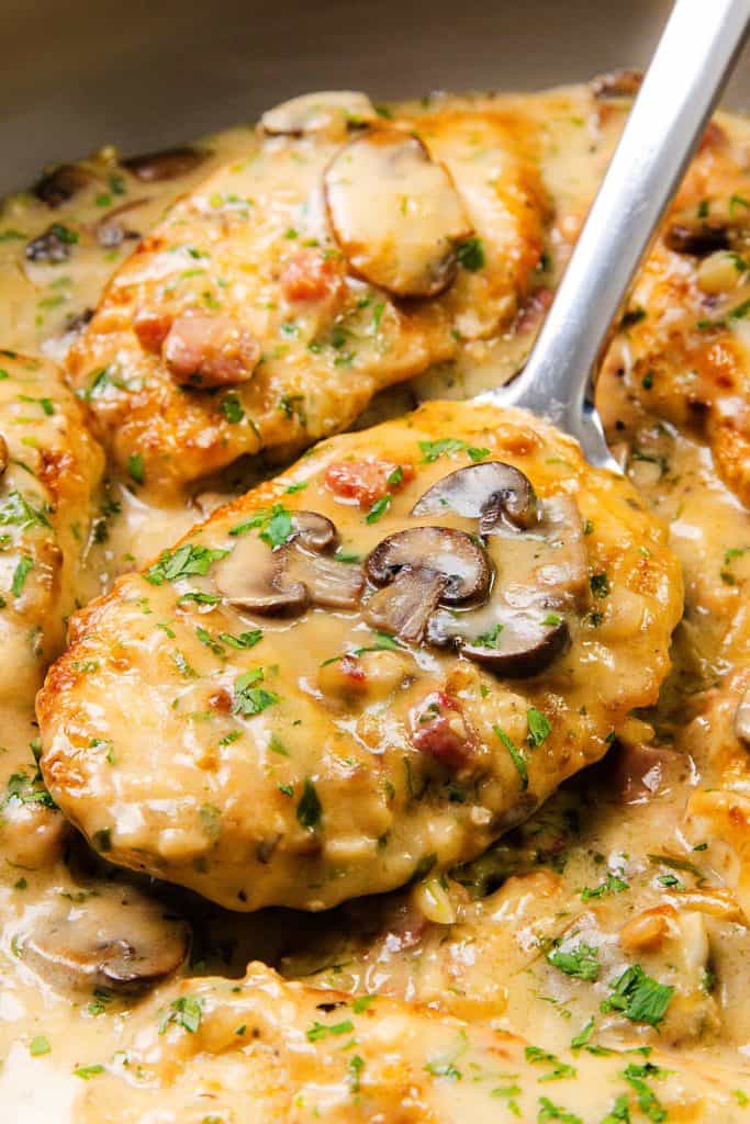 Chicken breasts in a creamy mushroom sauce, garnished with chopped herbs and bacon pieces. A spoon lifts one piece, revealing the rich, savory sauce and sliced mushrooms—perfect for fans of Creamy Chicken Marsala.