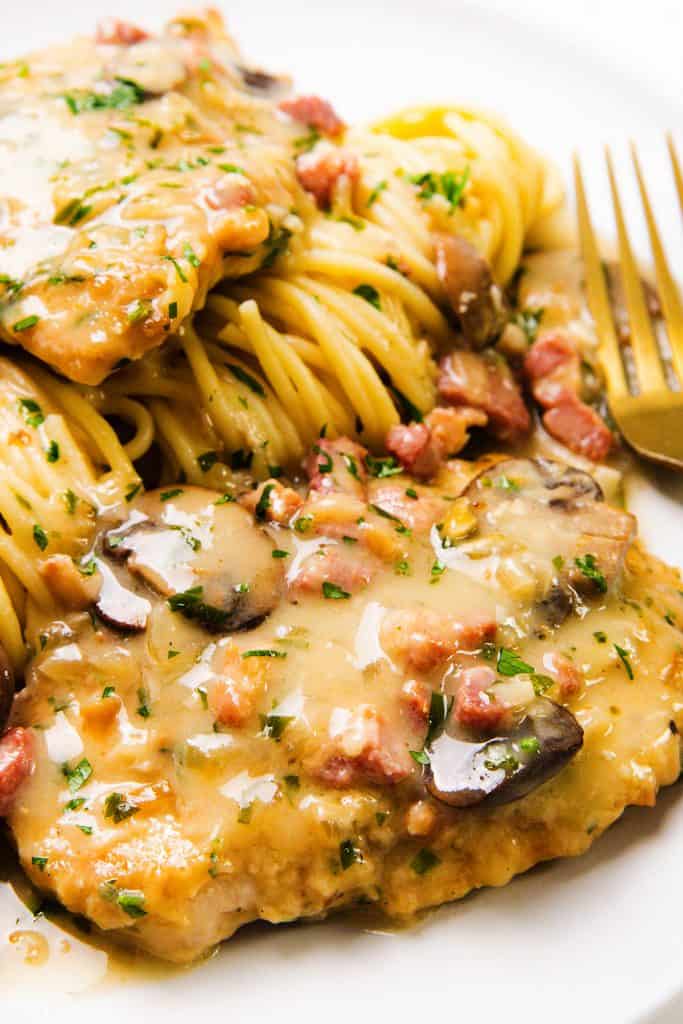 Close-up of Creamy Chicken Marsala with mushrooms and a rich sauce, served alongside pasta. A gold fork rests beside the food on a white plate.