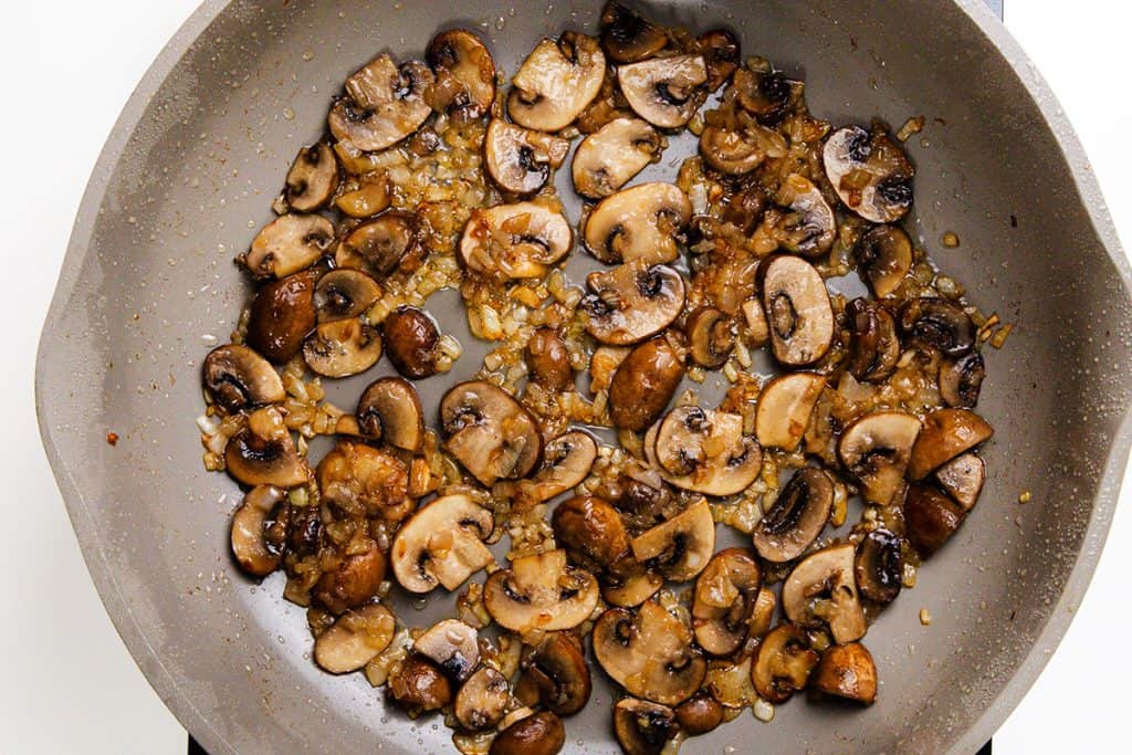 Sliced mushrooms and diced onions are being sautéed in a gray pan, turning golden brown and caramelized—the perfect base for a classic Chicken Marsala.