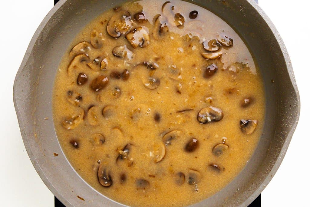 A gray skillet filled with rich, brown mushroom gravy reminiscent of Chicken Marsala, showing sliced mushrooms simmering in a thick, glossy sauce.