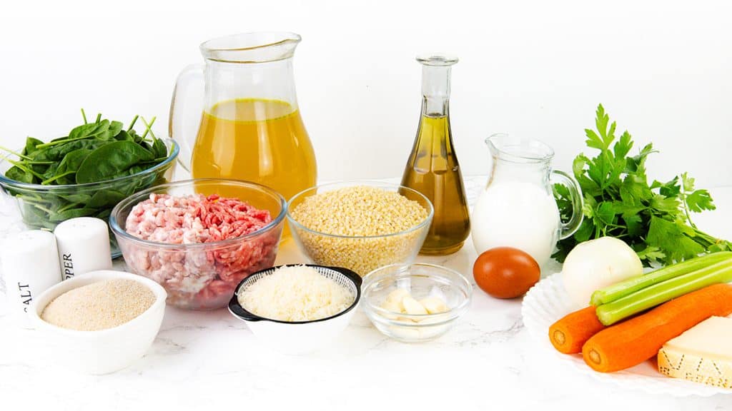 Ingredients for Italian Wedding Soup are arranged on a white surface, featuring ground meat, spinach, carrots, celery, onion, garlic, couscous, grated cheese, an egg, parsley, seasonings, olive oil, broth, milk, and breadcrumbs.