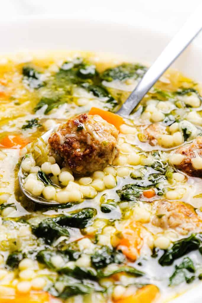 A close-up of Italian Wedding Soup in a bowl, featuring meatballs, small pasta, spinach, and carrots, with a spoon lifting a flavorful meatball and vegetables from the aromatic broth.
