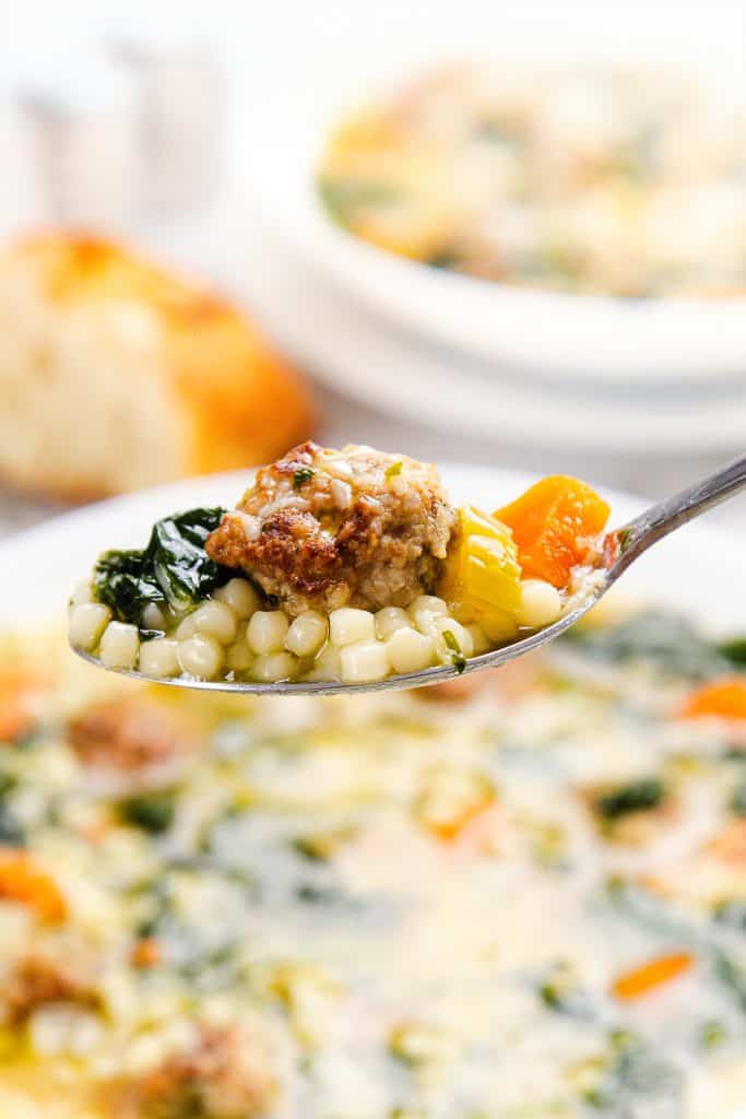 A close-up of a spoonful of Italian Wedding Soup, featuring mini meatballs, acini di pepe pasta, spinach, and diced carrots above a bowl of the classic Italian Wedding Soup, with bread blurred in the background.