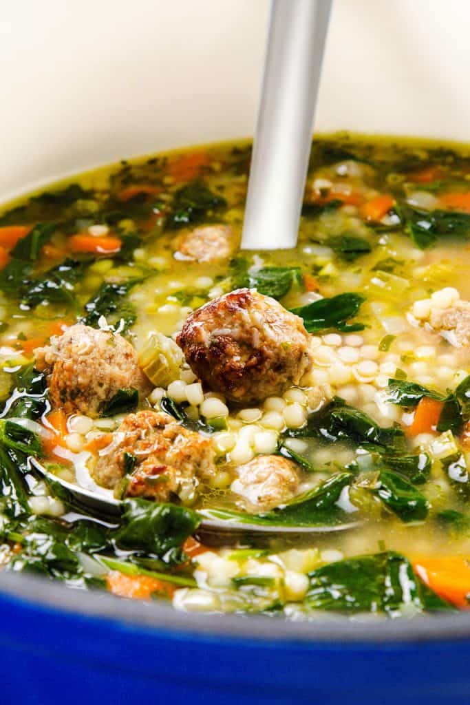 A close-up of Italian Wedding Soup with meatballs, carrots, leafy greens, pasta, and savory broth in a pot with a metal ladle.