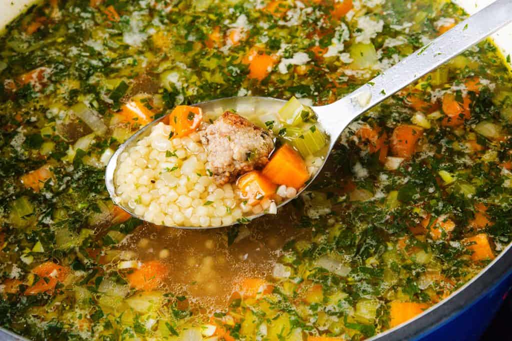 A close-up of a spoonful of Italian Wedding Soup with small pasta, carrots, celery, and herbs held above a pot brimming with colorful broth and diced vegetables.