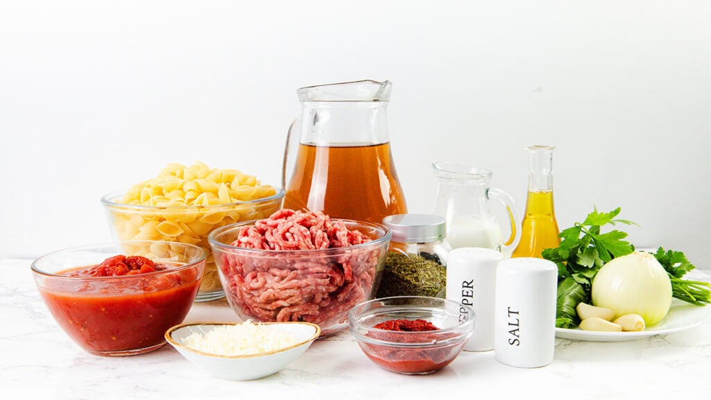 A variety of ingredients on a white surface, including bowls of pasta shells, ground meat, canned tomatoes, tomato paste, and shredded cheese—perfect for making a creamy ground beef pasta or delicious one pot pasta dish.