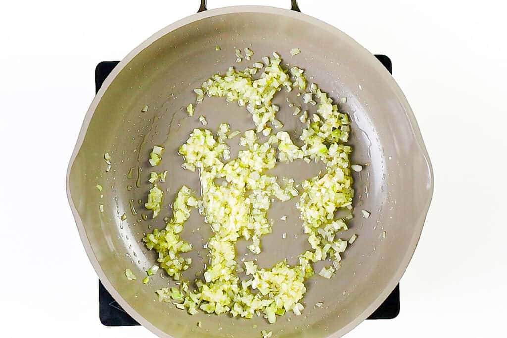 Chopped onions and garlic are being sautéed in a light-colored skillet on a stovetop, creating the flavorful base for a delicious creamy ground beef one pot pasta.