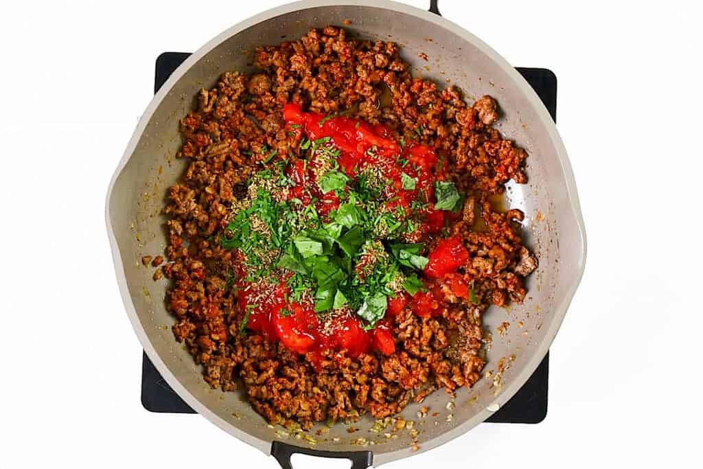 A skillet filled with creamy ground beef pasta, topped with tomato sauce, chopped fresh basil, and herbs, sits on a stovetop against a white background.