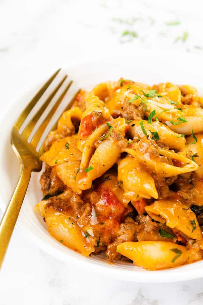 A white bowl filled with creamy ground beef pasta shells, mixed with cheese and garnished with chopped herbs, with a gold fork resting on the side.