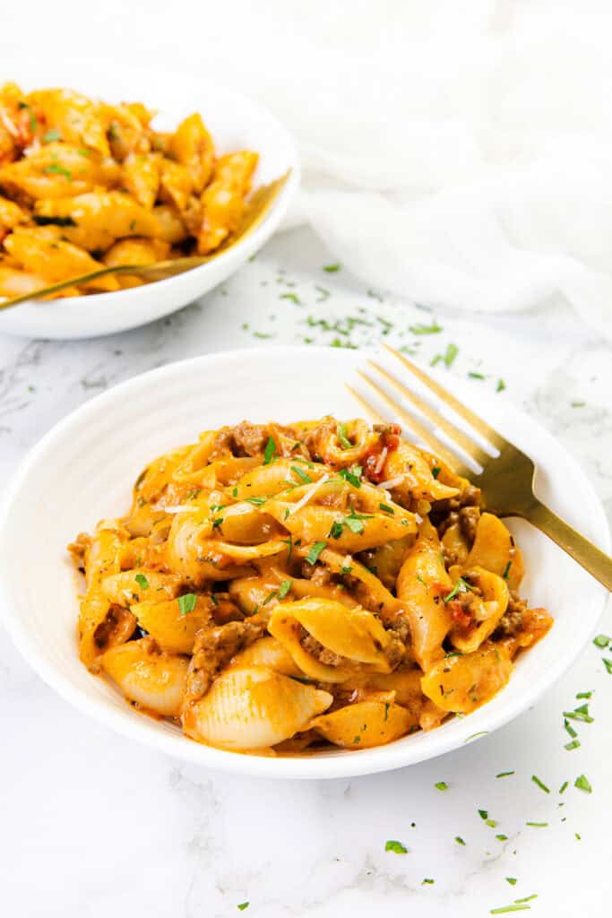 A white bowl filled with creamy tomato pasta shells mixed with ground beef and garnished with chopped parsley. A gold fork rests in the bowl, and another bowl of this delicious ground beef pasta sits on a white marble surface.