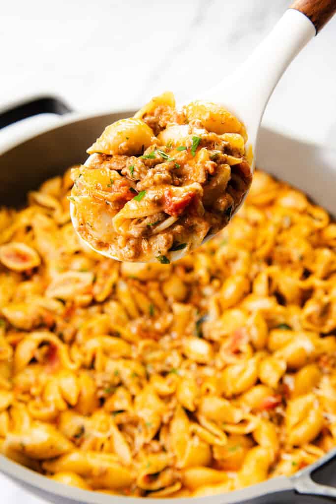 A close-up of a white spoon holding a serving of creamy ground beef pasta shells with herbs, above a pan filled with the same one pot pasta dish.