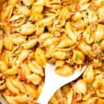 A close-up of a pan filled with creamy ground beef pasta shells in a rich sauce, mixed with herbs and grated cheese. A white serving spoon is scooping some of this hearty one pot pasta.
