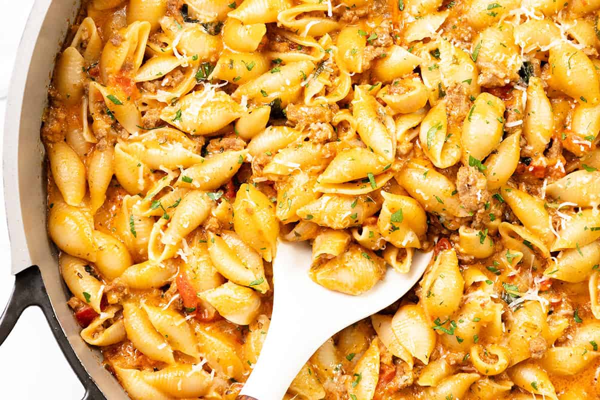 A close-up of a pan filled with creamy ground beef pasta shells in a rich sauce, mixed with herbs and grated cheese. A white serving spoon is scooping some of this hearty one pot pasta.