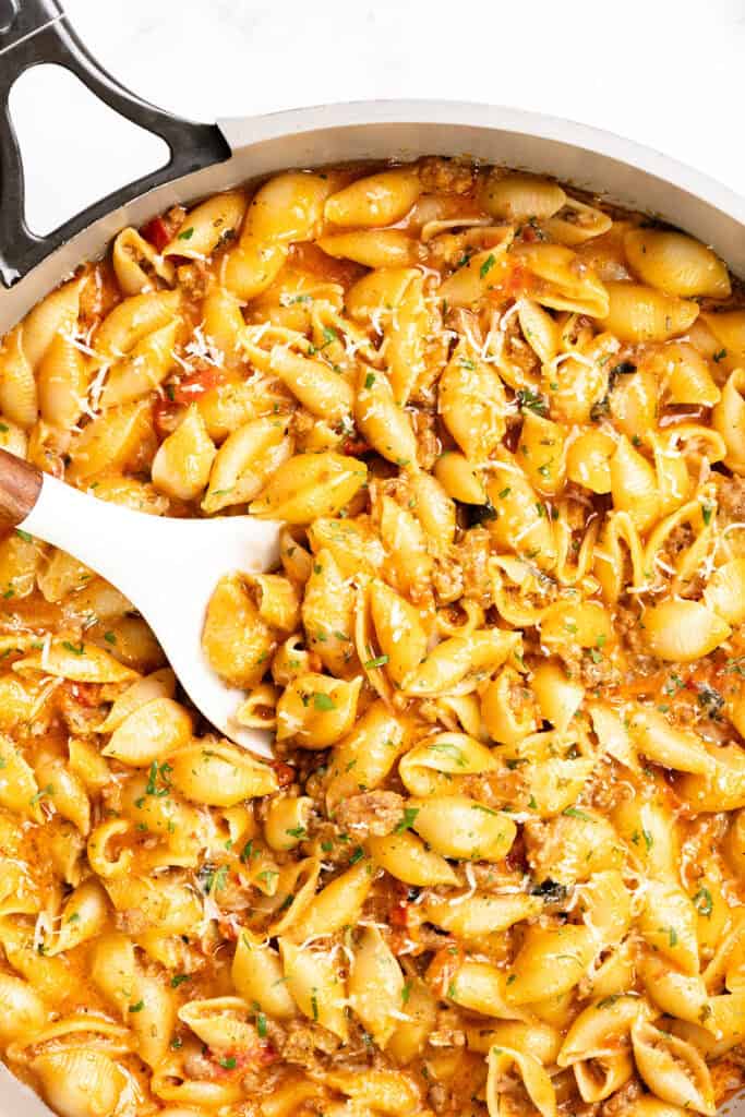 A large skillet filled with creamy tomato pasta shells in a rich orange sauce, garnished with herbs and shredded cheese, with a white serving spoon scooping some of this one pot pasta.