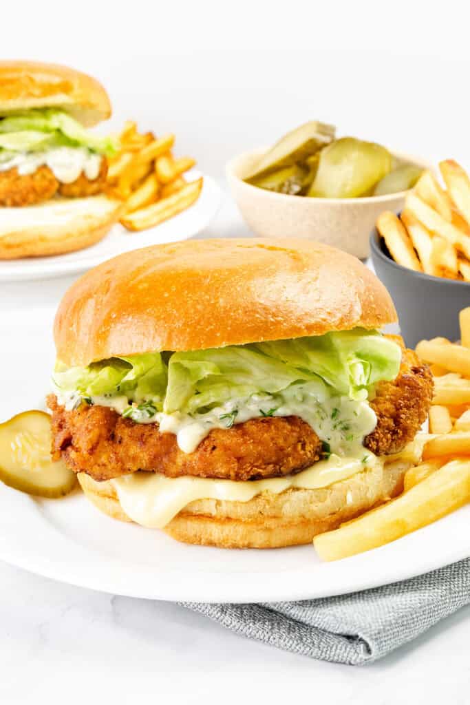 A crispy pickle brined fried chicken sandwich with lettuce, mayonnaise, and cheese on a bun, served with French fries and pickle slices. Another sandwich, extra fries, and a bowl of pickles are in the background.