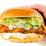 A hand holding a pickle brined fried chicken sandwich with lettuce, creamy sauce, and a golden toasted bun against a white background.