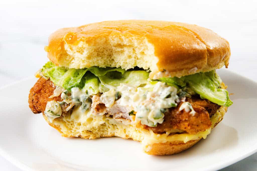 A close-up of a pickle brined fried chicken sandwich with a bite taken out, showing crispy chicken, lettuce, and creamy sauce inside a soft bun, served on a white plate.