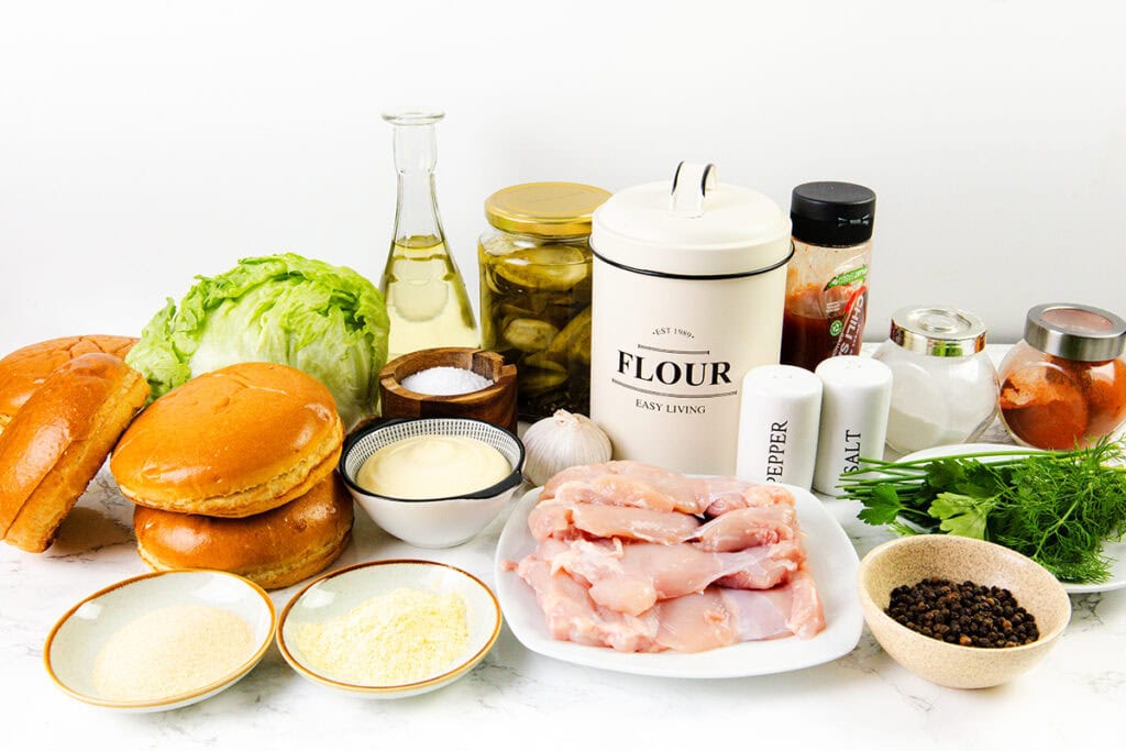 A variety of pickle brined fried chicken sandwich ingredients including raw chicken, buns, lettuce, flour, pickles, mayonnaise, oil, garlic, spices, salt, pepper, breadcrumbs, and fresh dill arranged on a white surface.