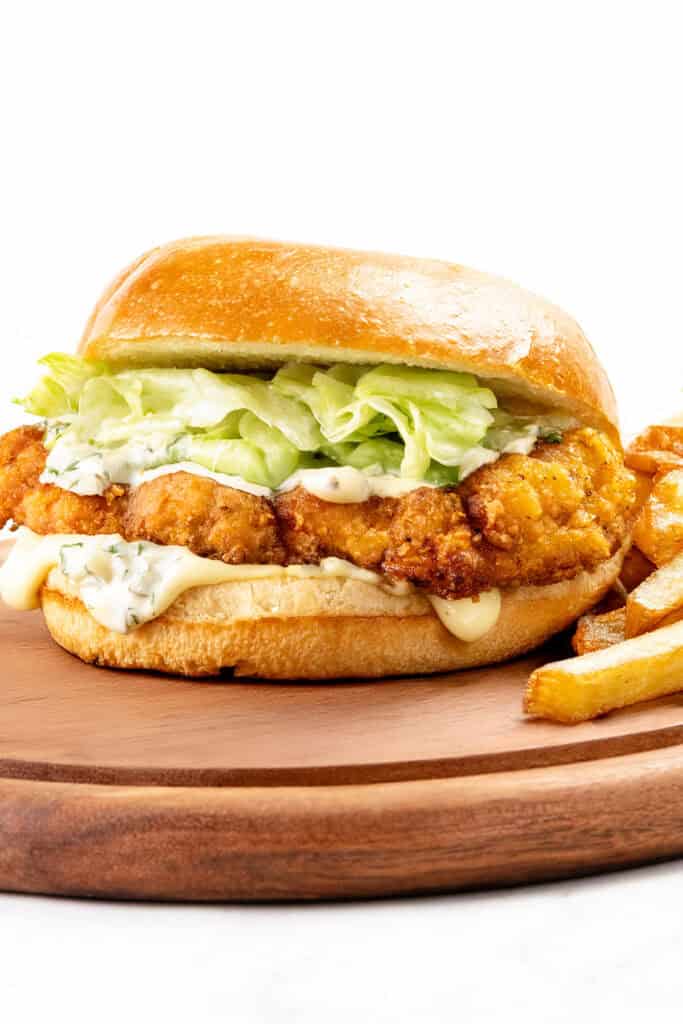 A crispy pickle brined chicken sandwich with lettuce and creamy sauce on a brioche bun, served on a wooden board with golden French fries on the side.
