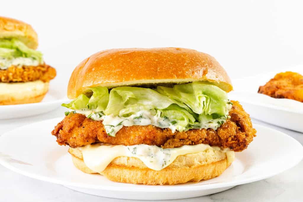 A crispy pickle brined fried chicken sandwich on a bun with mayonnaise, lettuce, and creamy sauce, served on a white plate with another sandwich and a piece of fried chicken in the background.