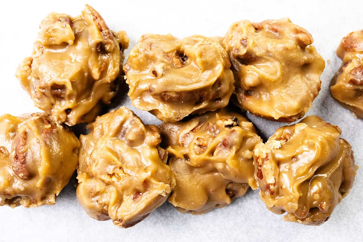 Close-up of several clusters of creamy, golden-brown Southern Pralines candy on a white surface, showcasing their rough texture and visible pecan pieces.