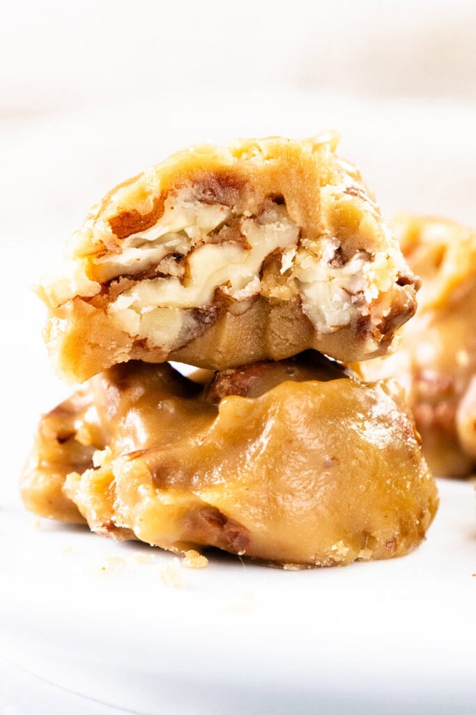 Two pieces of creamy Southern Pralines candy with a golden caramel coating, one broken open to reveal chopped pecans inside, stacked on a white surface.