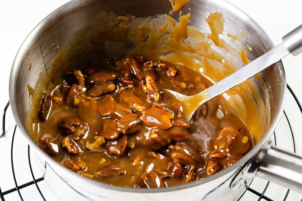 A saucepan filled with glossy caramel and pecans, perfect for Southern Pralines, is being stirred with a metal spoon on the stove, caramel splatters decorating the sides as this classic Southern candy comes together.